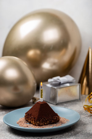 Small chocolate cake with burning candle against the background of golden balloons and gift box. Birthday celebration conceptの写真素材