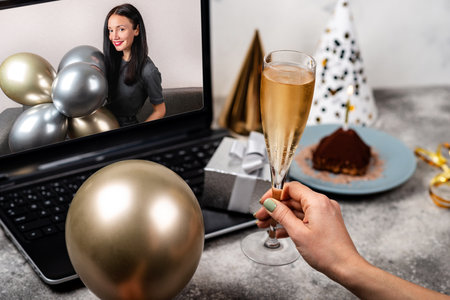 Birthday party in online video call. Female hands are holding glass of wine and balloon in front of laptop screen. Young woman celebrating her birthday at home. Remote Celebration Concept.の写真素材