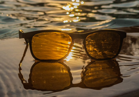 Stylish sunglasses resting gently on wet surface glowing with golden sunset hues and delicate watery reflectionsの写真素材