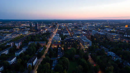 An aerial shot captures a city at twilight.  The sky displays soft hues of blue and pink.  Numerous buildings of varying heights and architectural styles are visible, many with illuminated windows. A main street cuts through the scene, showing light trails from moving vehicles.  Surrounding the urban area are stretches of dark green trees. The overall mood is serene and peaceful, conveying a sense of calm within a bustling city environment. The image is sharply focused, offering a detailed view of the city's layout and infrastructure.の写真素材