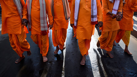A high-angle, full shot depicts several men in saffron-colored robes walking barefoot on wet asphalt.  The men are walking in a line, their shadows cast long on the dark, glistening surface. The robes are a deep, rich orange, and some have white scarves draped around their necks. The overall mood is somber and peaceful, with a focus on the repetitive pattern of the walking figures and their shared attire. The wet asphalt reflects some light, creating a contrast with the dark shadows. The image has a slightly desaturated color palette, emphasizing the texture of the wet asphalt and the folds of the robes.の写真素材