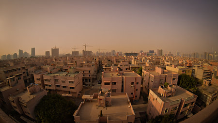 A high-angle, wide shot captures a cityscape dominated by numerous beige and tan buildings.  The buildings are primarily multi-story residential structures with flat roofs.  A hazy, dusty atmosphere gives the sky a muted, warm tone, suggestive of either sunset or a generally dusty environment.  Several construction cranes are visible in the distance, indicating ongoing development. The overall mood is calm and somewhat muted due to the color palette and atmospheric conditions. The image offers a detailed view of the rooftops and the dense arrangement of buildings, extending to a skyline of taller structures in the background.の写真素材