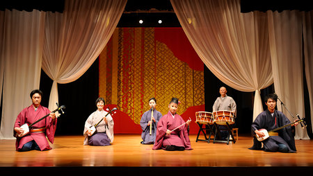 Six Japanese musicians are seated on a wooden stage performing traditional music.  Two play the shamisen, one plays the shakuhachi flute, one plays the taiko drum, and two more play the shamisen. They wear kimonos in shades of red, purple, and beige. The backdrop features a large red panel with a gold geometric pattern, framed by flowing beige curtains. The overall mood is serene and traditional. The image is well-lit and sharply focused, showcasing the detail of the instruments and clothing.の写真素材