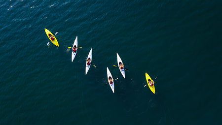 An aerial shot depicts six kayakers paddling on dark blue water. Two kayaks are yellow, and four are white. The kayakers are positioned in a slightly staggered line, suggesting a group activity. The water appears calm, and the overall mood is serene and active. The image is sharply focused, with clear visibility of the kayaks and paddlers against the deep blue backdrop. The high-angle perspective provides a unique view of the kayakers and their surroundings.の写真素材