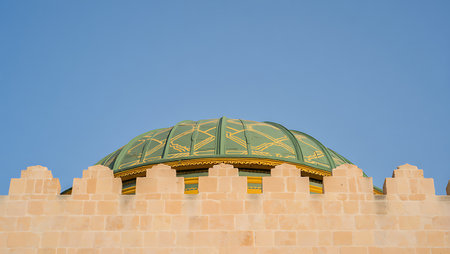 Close-up view of a building's architectural detail.  A green dome with a gold geometric pattern is prominently featured against a clear blue sky. The dome sits atop a tan or beige stone wall with a crenellated top. The stonework appears smooth and the colors are muted and warm. The image is sharply focused and the composition is simple, emphasizing the architectural elements. The overall mood is serene and peaceful.の写真素材