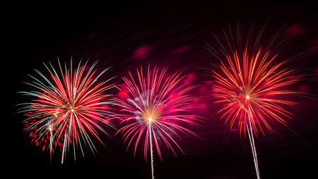 Three vibrant fireworks bursts illuminate a dark night sky. The central firework is predominantly pink and red, while the firework on the left features red and white streaks, and the firework on the right is primarily red with some orange highlights.  The fireworks are sharply defined against the black background, creating a striking contrast. The overall mood is celebratory and festive. The image is well-composed, with the fireworks arranged in a pleasing visual balance.の写真素材