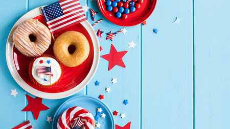 A high-angle, close-up shot depicts a vibrant arrangement of Fourth of July themed desserts and decorations on a light blue wooden surface. Two glazed donuts, one with a tan glaze and the other a plain yellow, sit on a red and white plate alongside a cupcake topped with white frosting and a miniature American flag. A red bowl filled with red and blue candies is also visible, along with scattered red, white, and blue star-shaped confetti.  The overall color palette is bright and cheerful, reflecting the festive nature of the scene. The image is well-lit and the textures of the food and decorations are clearly visible.の写真素材