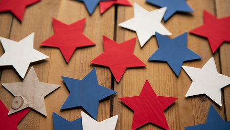 The image shows numerous five-pointed stars in red, white, and blue arranged on a light brown wooden surface.  The stars appear to be made of a painted material with a slightly textured appearance. They are scattered across the wood grain, creating a visually appealing and somewhat random pattern. The colors are vibrant and the overall mood is festive and patriotic, suggesting a connection to American holidays or celebrations. The wood grain is visible between the stars, providing a natural contrast to the bright colors.の写真素材