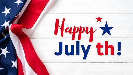 A folded American flag is positioned on a white wooden surface.  The flag's red, white, and blue colors are prominent.  Overlaid on the wood is the text "Happy July th!" in red and blue lettering, accompanied by small red and blue stars. The image is a flat lay composition, with a clean and simple style. The wood grain is visible, providing a textured contrast to the smooth fabric of the flag. The overall mood is celebratory and patriotic.の写真素材