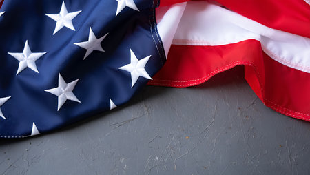 A close-up shot of a portion of an American flag draped on a gray textured surface. The flag's deep blue field with white stars is prominently featured, along with a section of the red and white stripes. The gray background has a slightly rough, concrete-like texture, providing a contrast to the smooth fabric of the flag. The image is composed with the flag taking up a significant portion of the frame, leaving ample space on the gray background. The overall mood is one of solemnity and patriotism. The lighting is even and soft, highlighting the details of the flag's fabric and the texture of the background.の写真素材