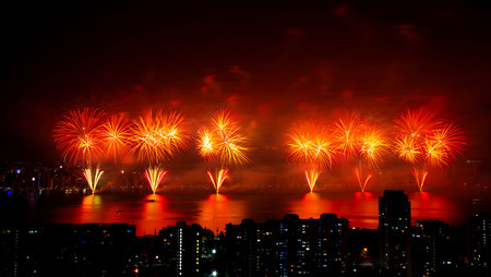 A nighttime long shot of a vibrant fireworks display over a city skyline.  The fireworks are predominantly orange and red, creating a fiery spectacle against the dark night sky.  The explosions are large and numerous, reflected in the dark water below. The city buildings are silhouetted against the bright fireworks, adding to the dramatic effect. The overall mood is celebratory and energetic. The image is sharp and well-exposed, capturing the detail of both the fireworks and the city.の写真素材
