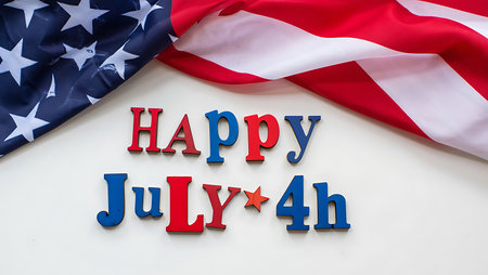 A high-angle, close-up shot depicts the phrase Happy July 4th spelled out in red, white, and blue wooden letters against a white background. A portion of an American flag is draped diagonally across the upper right corner of the image. The letters are arranged neatly, and the flag's folds create a textured contrast to the smooth white surface. The overall mood is celebratory and patriotic, evoking the spirit of Independence Day. The color palette is limited to the classic red, white, and blue of the American flag, creating a clean and visually appealing composition.の写真素材