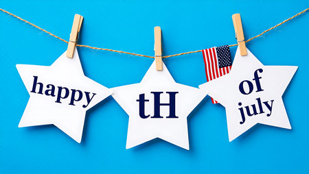 Three white paper stars are strung together on a twine line against a bright blue background. Each star has navy blue text: the first says "happy", the second "th", and the third "of july". A small American flag hangs between the second and third stars.  The stars are attached with small wooden clothespins. The overall mood is celebratory and festive.の写真素材