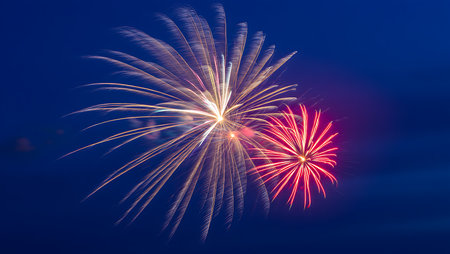 A photograph of a fireworks display at night. Two bursts of fireworks are visible against a deep blue sky. One burst is predominantly white and gold, while the other is a vibrant red. The fireworks create a dramatic and colorful spectacle in the night sky. The image is sharp and well-exposed, capturing the intricate details of the fireworks bursts. The overall mood is celebratory and festive.の写真素材