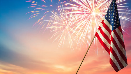 A photograph depicts an American flag waving gently on a wooden pole against a backdrop of a vibrant sunset and a dazzling fireworks display. The flag's red, white, and blue colors are prominent, and the fireworks create a burst of light in shades of white, pink, and orange. The sky transitions from a soft blue at the top to warm orange and pink hues near the horizon. The overall mood is celebratory and patriotic. The image is well-composed, with the flag positioned to the right, allowing ample space for the fireworks and sky.の写真素材