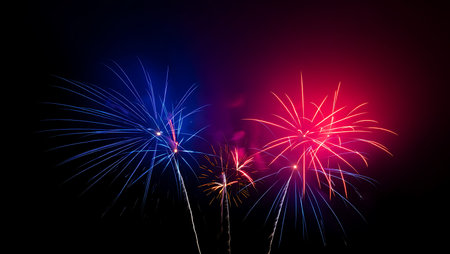 Three fireworks bursts illuminate the night sky. One firework is predominantly blue, another is...
