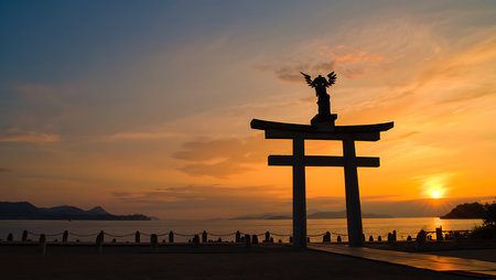 Orange and yellow sunset over calm ocean waters. Silhouetted torii gate with angel statue stands in foreground. Mountains visible in distance. Peaceful, serene mood.の写真素材