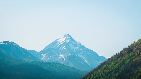 A snowcapped mountain peak dominates the center of the image, flanked by verdant forested hills under a pale blue sky.  The colors are muted and cool, creating a serene atmosphere.の写真素材