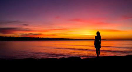 Silhouette of a young woman standing on a rock by the sea at sunsetの写真素材
