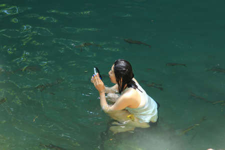 girl in a bikini is standing in emerald green water and taking a picture of herself with a mobile phone in a waterfall with a swarm of fish swimming nearby.のeditorial素材