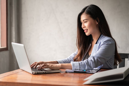 Young happy Asian businesswoman in blue shirt working from home and use a computer laptop and thinking idea for her businessの写真素材