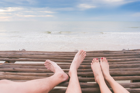 The couple's legs sit on wood bridge overlooking beautiful sea.の写真素材
