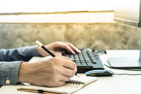 Close up of Male hands writing some data in notebook and using computer on the deskの写真素材