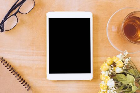 Top view of tablet with tea cup, flower, notebook and glasses on wooden tableの写真素材