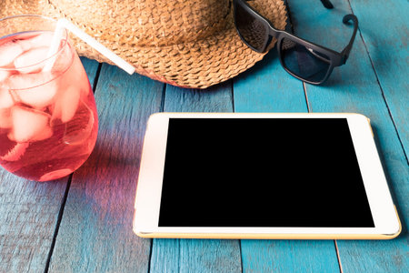 Tablet with hat, sunglasses and red cocktail on blue painted wood plank background.