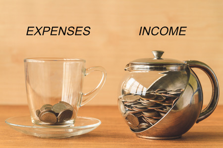 Coins in a cup and a jar on a wooden table, expenses and income text, financial conceptの写真素材
