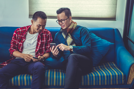 Two friends talking and using smart phone while sitting on sofa at home.の写真素材