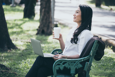 Asian businesswoman working on laptop while sitting on the bench and relaxing with a cup of coffee in a public park.の写真素材