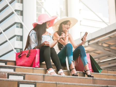 Two beautiful young women sitting on the stairs and using smart phone after enjoying in shoppingの写真素材