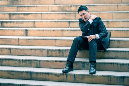 Asian business man sitting on city stairs with feeling pain in his shoulder.の写真素材