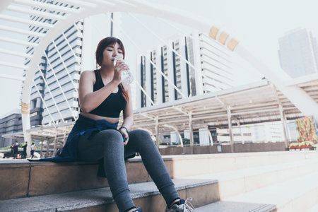Young Asian woman is relaxing and drinking water after jogging in city.