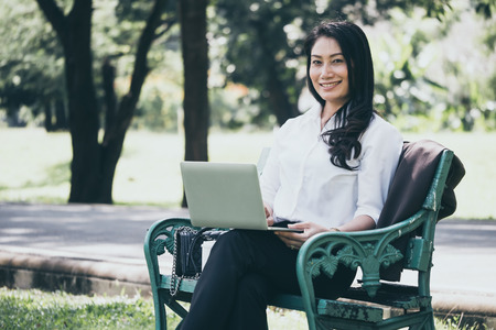 Happy Asian businesswoman working on laptop while sitting on the bench in the parkの写真素材