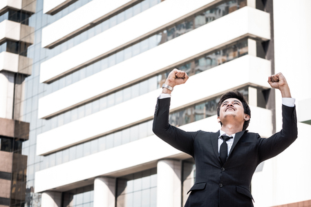 Portrait of young Asian businessman in black suit standing with office building background. Free space for textの写真素材