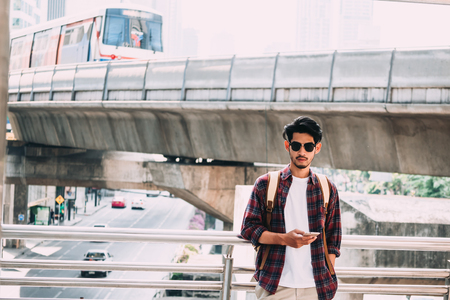 Portrait of young asian man in casual clothes with sunglasses, smart phone and backpack on urban scene background.の写真素材