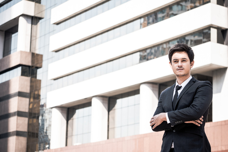 Portrait of young Asian businessman in black suit standing with office building background. Free space for textの写真素材