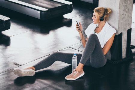 Young happy woman sitting and listening to music by smartphone while having exercise break at the...