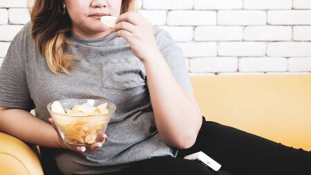Chubby woman eating potato chips and sitting on a sofa in a room.の写真素材