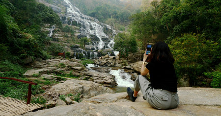 Woman in black t-shirt sitting and taking photos of beautiful waterfall by smartphone.の写真素材