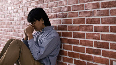 Christian Asian man is sitting against a wall, feeling sad, holding a cross and praying to God.の写真素材