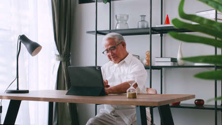 An elderly man reading news with the tablet on the desk at home. An old Asian man is searching for information on the Internet while sitting at a table.の写真素材