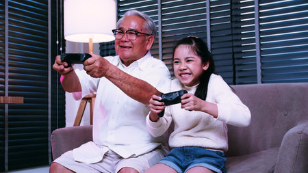 Grandpa and granddaughter are having fun playing video games together in the living room at home.