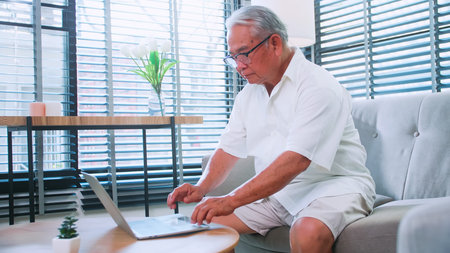 An elderly man reading news with the tablet on sofa at home. An old Asian man is searching for...