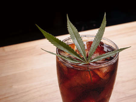 Glass of cola with ice cubes and cannabis leaf on the wooden table.の写真素材