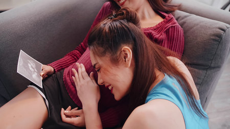 Happy pregnant lesbian couple looking ultrasound image together on sofa.