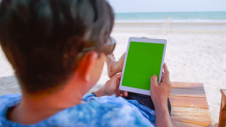 Asian man watching something on his tablet while relaxing on the beach.