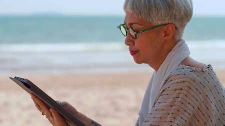 Asian elderly woman using a tablet computer while sitting on the beach.の写真素材
